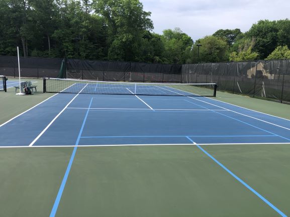 Regulation outdoor tennis court surrounded by black fence with trees in background